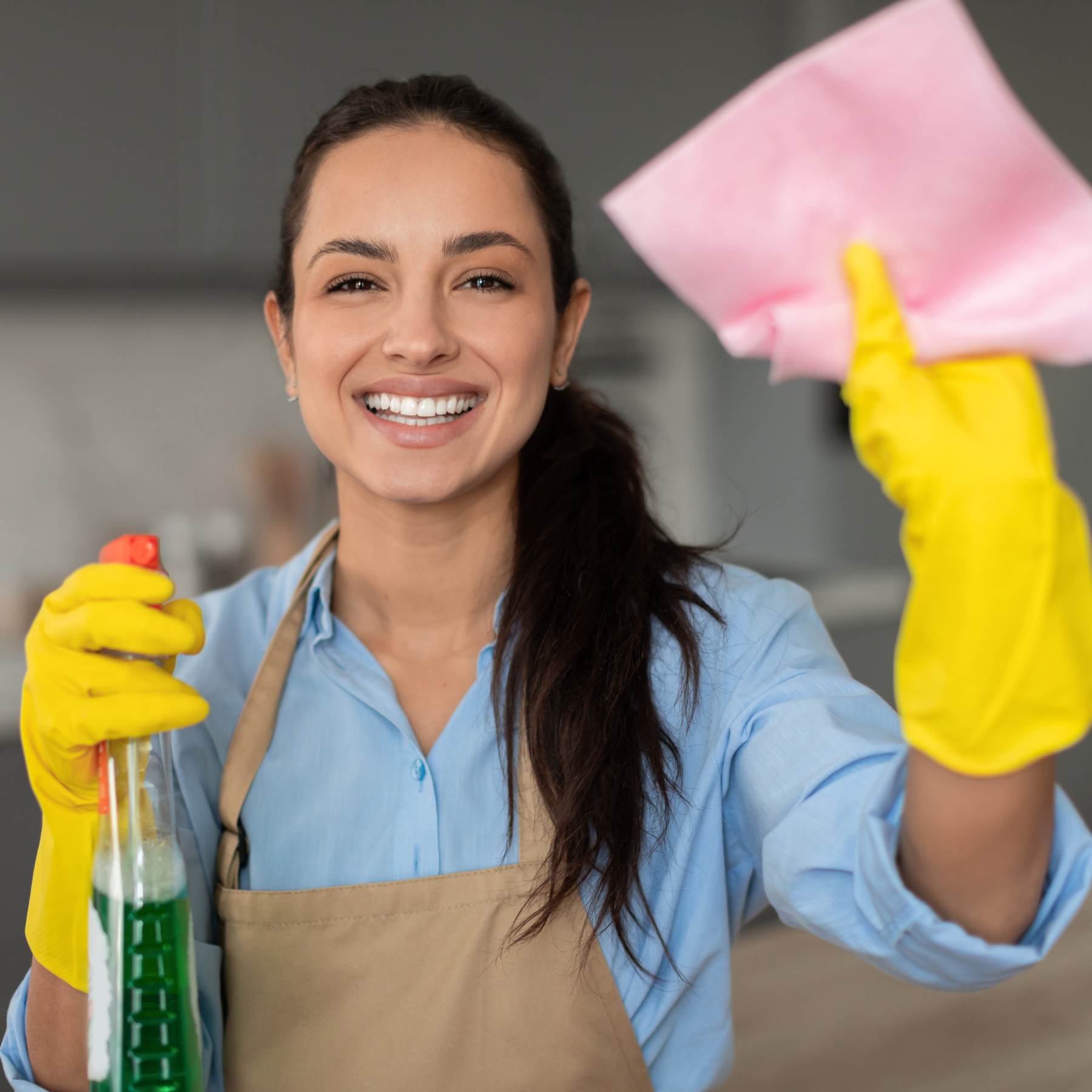 Femme en tablier tenant un chiffon et des produits de nettoyage, prête à faire le ménage.